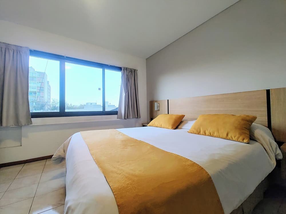 Habitación con cama grande, dos almohadas, ventana con cortinas y edificios