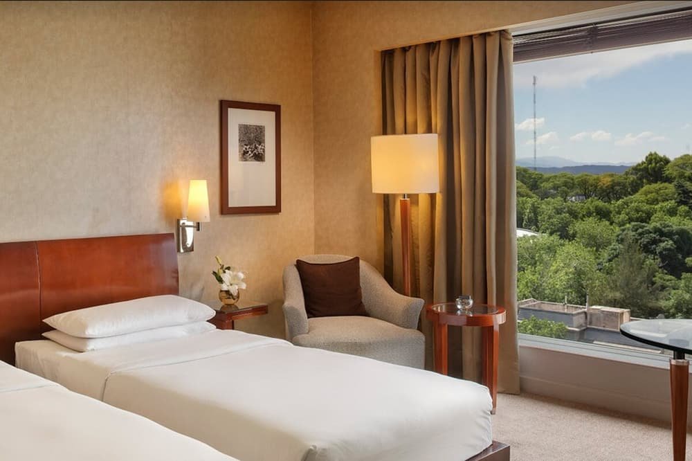 Quarto com duas camas, poltrona, mesa e janela com vista para árvores e montanhas