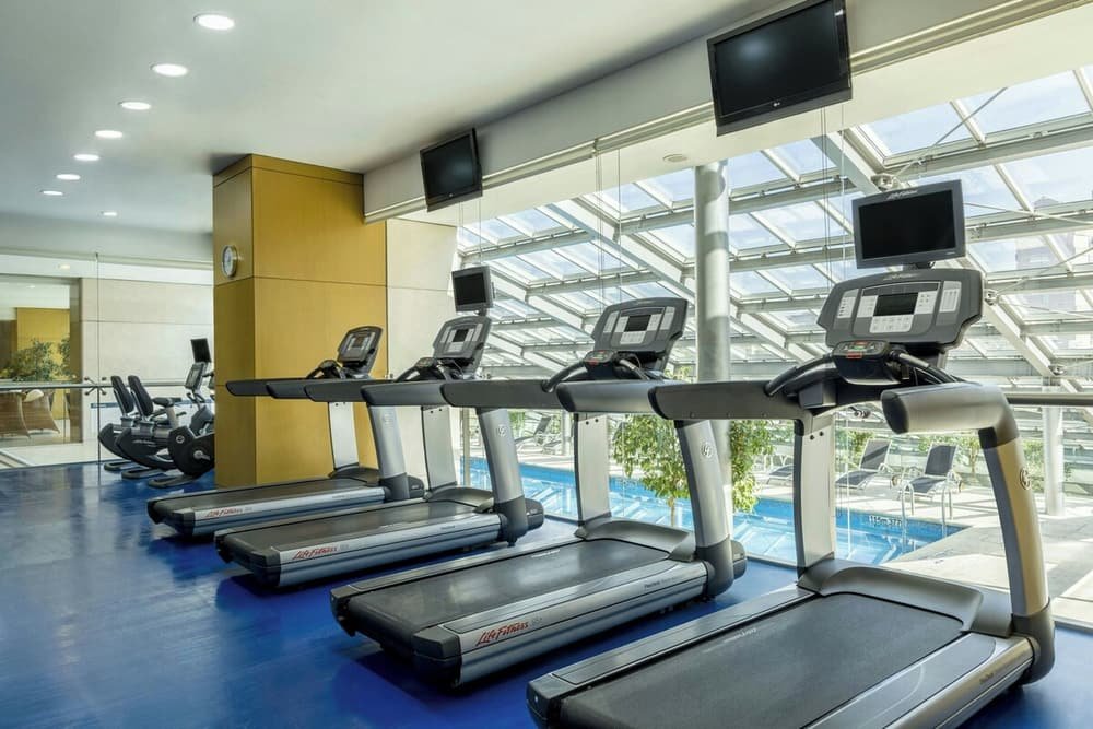 Academia com esteiras e bicicletas ergométricas, com a área da piscina ao fundo